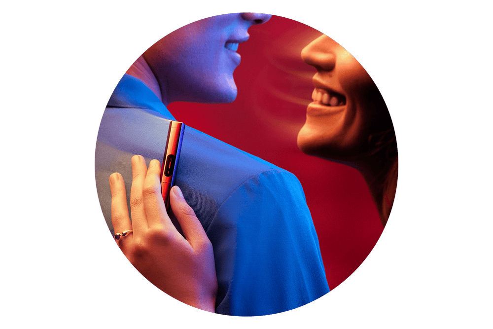 A woman smiles while holding a sleek, colorful vape device against a mans shoulder, both lit with vibrant blue and red lighting.