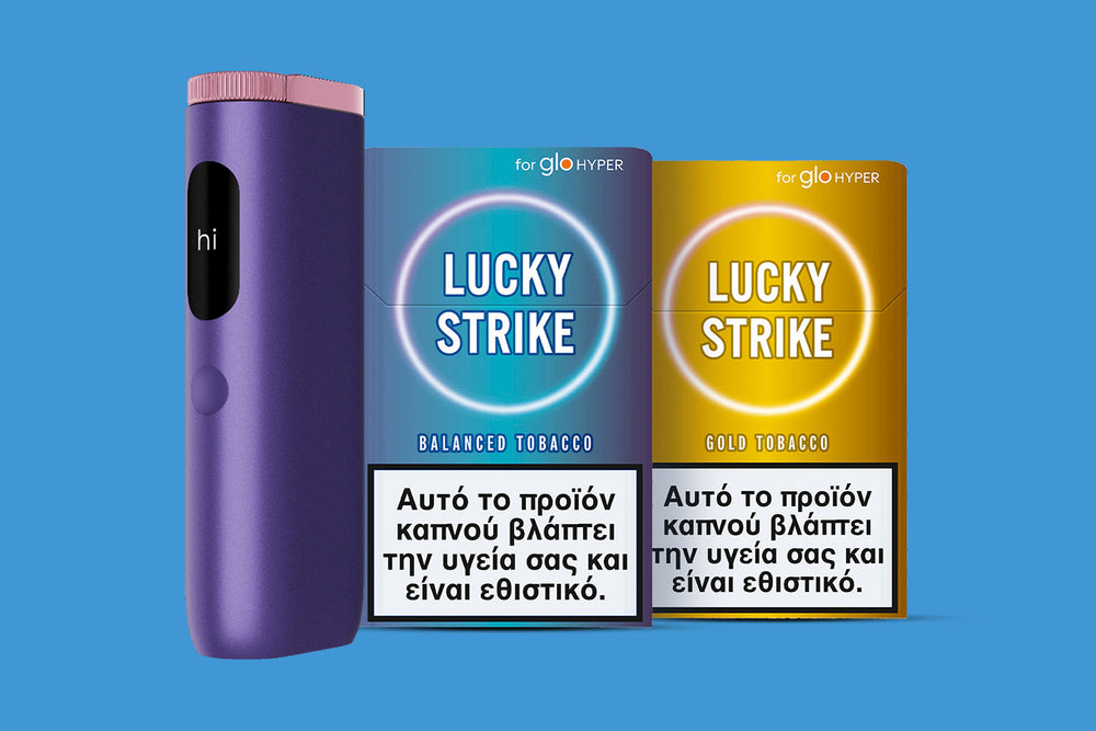 A purple electronic device labeled hi stands next to two Lucky Strike tobacco product packs with Greek health warnings on a blue background. One pack is blue (Balanced Tobacco) and the other is yellow (Gold Tobacco).