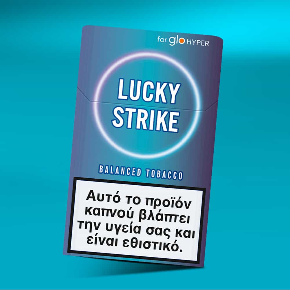 A Lucky Strike Balanced Tobacco blue cigarette pack with a Greek health warning at the bottom sits on a turquoise surface against a blue background, emphasizing its mid-intensity, warm, tobacco-dominant blend.