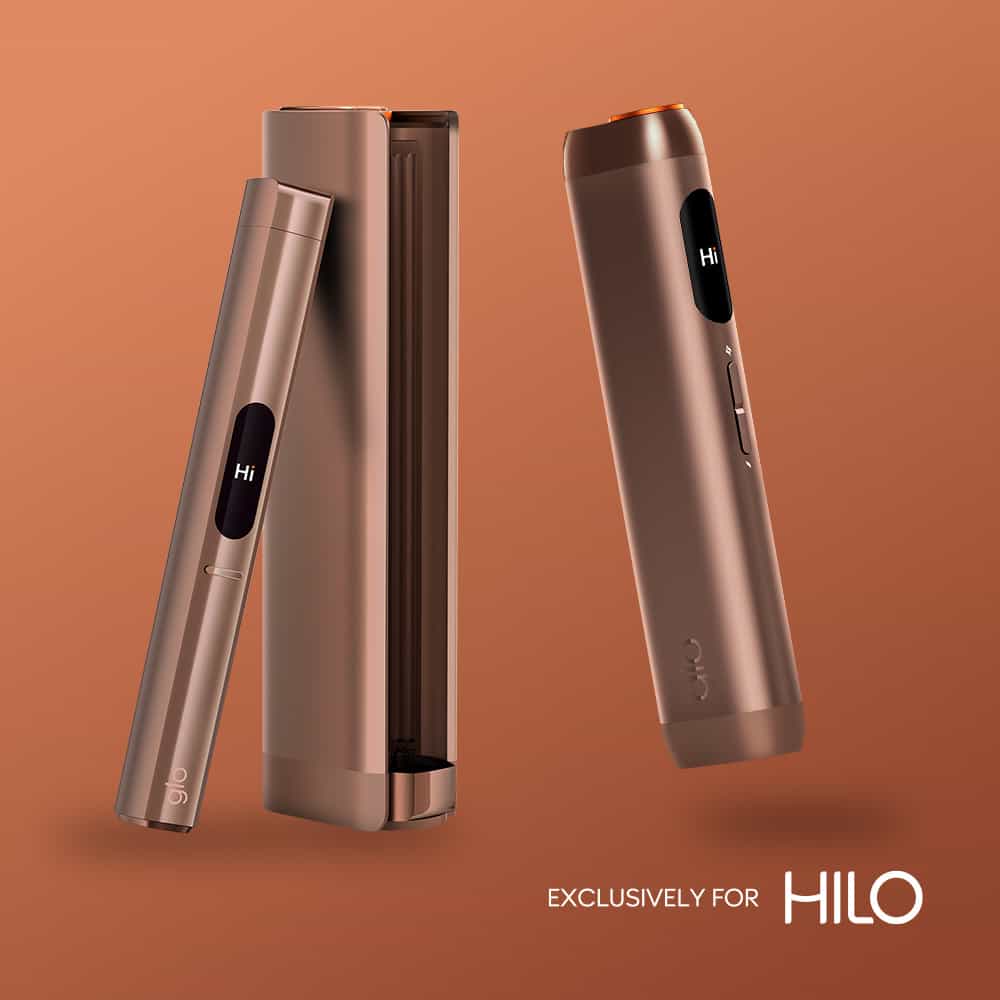 Two sleek, metallic bronze virto™ Golden Tobacco devices with small screens displaying Hi stand against a matching brown background. One device is in its charging case. Text reads Exclusively for HILO.