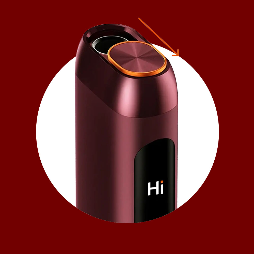 A close-up of a maroon-colored electronic device with a round, metallic orange button on top, an orange arrow pointing to the button, and the word Hi displayed on a black screen.
