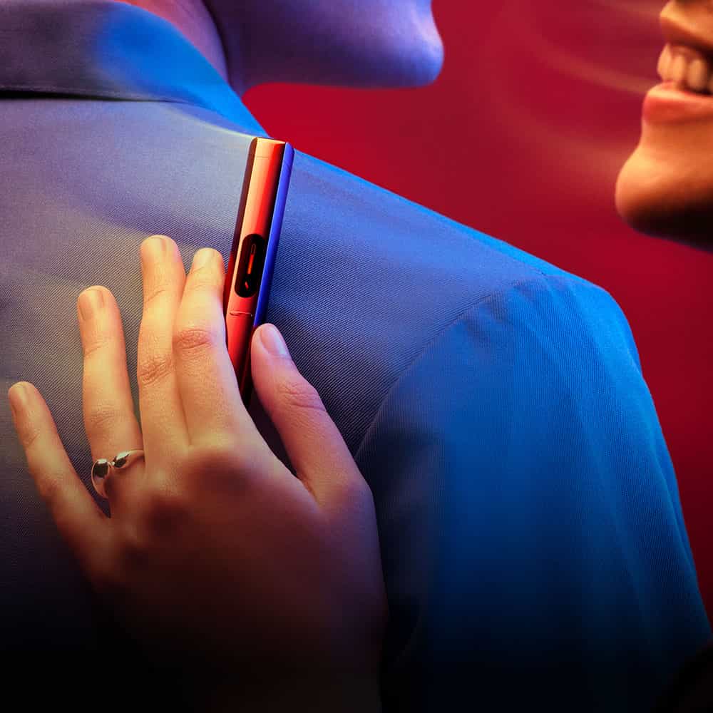 A close-up of a hand holding a red vape pen against someones shoulder. Two people are smiling, one with a visible engagement ring, in a warmly lit, red and blue background.